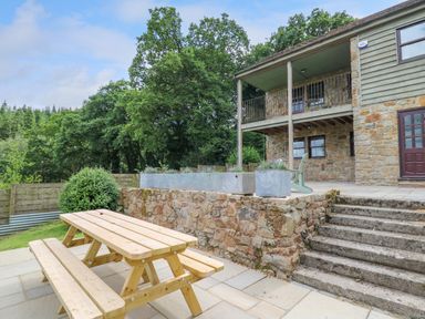 An outdoor patio area with a wooden picnic table and stone steps leading to a two-story stone house with a covered balcony and potted plants at Oak Apple Cottage in Presteigne