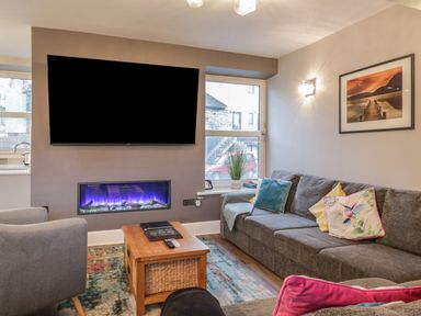 A living room with a gray sofa and colorful cushions a wooden coffee table a wall-mounted television and an electric fireplace at Quarters in Ambleside