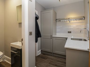 A utility room with grey cabinets a countertop and sink a coat hanging and a small adjacent bathroom at Quarters Ambleside