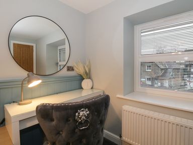 A desk with a lamp a vase and a round mirror above a chair next to a window with blinds at Quarters in Ambleside
