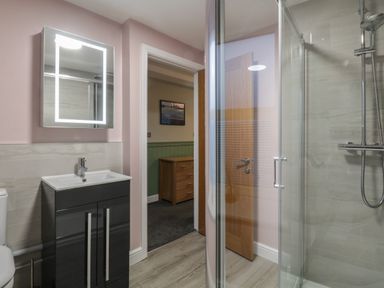 A bathroom with a sink and cabinet a glass shower enclosure and a door leading to a room with a chest of drawers at Quarters in Ambleside