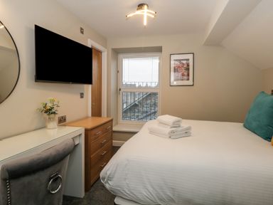 A bedroom with a bed with towels folded on it a window with blinds a wooden chest of drawers a wall-mounted tv and a desk with a chair and flowers at Quarters in Ambleside