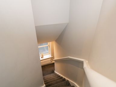 A staircase with carpeted steps and white walls leading down to a window with a radiator below at Quarters in Ambleside