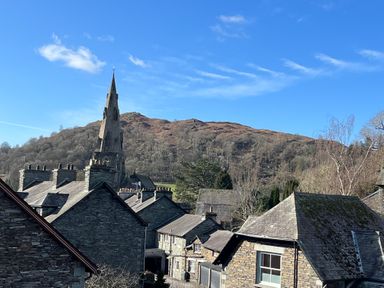 Stone buildings with slate roofs and a tall church spire with a hill and blue sky in the background at Quarters Ambleside