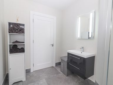 A bathroom with a white door a cabinet with rolled towels a sink with vanity drawers a lighted mirror and a laundry hamper at Curhoor Near Loughrea County Galway