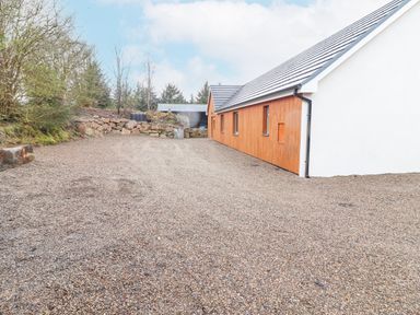 A gravel driveway beside a modern building with wooden siding and white walls near a stone retaining wall with trees at Curhoor Near Loughrea County Galway