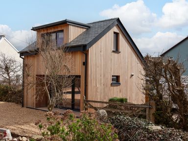 A wooden house with black trim and a gravel yard with leafless trees at The Cabin at Ropewalk in Goonbell near St Agnes