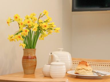 A table with a vase of yellow flowers a white teapot a sugar bowl and a plate with a scone at The Cabin at Ropewalk in Goonbell near St Agnes