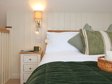 A bedroom with a bed with white sheets and green pillows a bedside table with books and a mug and a wall lamp at The Cabin at Ropewalk in Goonbell near St Agnes