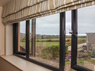 A window with striped blinds partially open showing a rural view with fields bushes and a road at The Cabin at Ropewalk in Goonbell near St Agnes