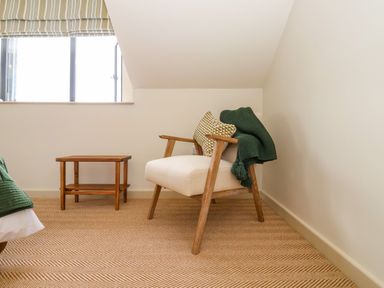 A corner of a room with a wooden armchair with cushions and a throw and a small wooden side table at The Cabin at Ropewalk Goonbell near St Agnes