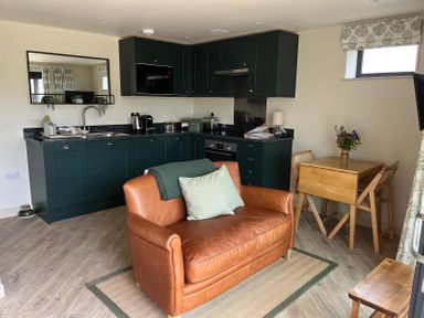 A kitchen with dark green cabinets a small brown leather sofa and a wooden dining table with chairs and flowers at The Cabin at Ropewalk in Goonbell near St Agnes