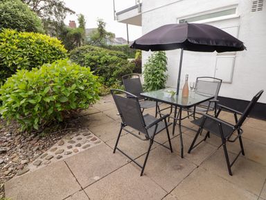 A patio with a glass table and four chairs under a black umbrella next to green bushes at 14 Coedrath Park in Saundersfoot