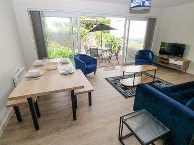A living room with blue sofas and chairs a wooden dining table set with dishes and glassware and a TV on a stand at 29 Coedrath Park Saundersfoot