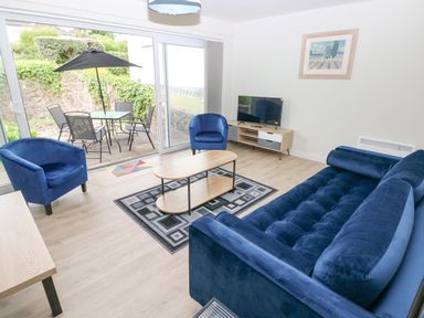 A living room with blue sofa and chairs a coffee table and television with sliding glass doors to a patio at 29 Coedrath Park Saundersfoot