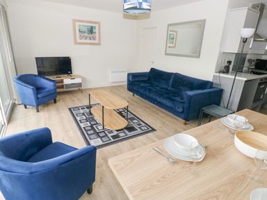A living room with blue armchairs and sofa a wooden coffee table a TV on a stand and a dining table with plates and glasses at 29 Coedrath Park in Saundersfoot