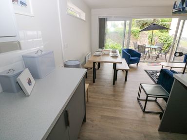 A dining area with a wooden table set for four and blue chairs next to glass sliding doors leading to an outdoor patio with a table and umbrella at 29 Coedrath Park in Saundersfoot
