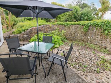An outdoor patio with a glass table and black chairs under a large black umbrella surrounded by a stone wall and greenery at 29 Coedrath Park in Saundersfoot