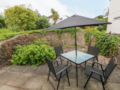 An outdoor patio area with a glass table and four chairs under a large black umbrella surrounded by plants and a stone wall at 29 Coedrath Park in Saundersfoot