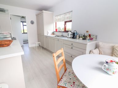 A kitchen with white cabinets a wooden chair with floral cushion a white round table and various kitchen appliances at Tonakilla House in Tonakilly near Camp County Kerry