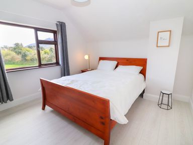A bedroom with a wooden bed frame white bedding a bedside table with a lamp a window with gray curtains and a small stool near a wall at Tonakilla House Tonakilly near Camp County Kerry