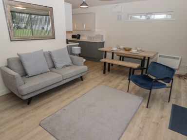 A living area with a gray sofa, a blue armchair, a wooden dining table with bench seats, and a kitchen in the background at 23 Coedrath Park Saundersfoot