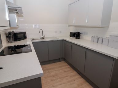 A kitchen with gray lower cabinets white upper cabinets a sink a black stovetop a microwave a toaster and a kettle at 23 Coedrath Park Saundersfoot