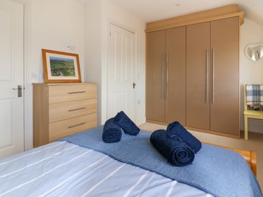 A bedroom with a bed with rolled towels on a blue blanket a chest of drawers with a framed picture a brown wardrobe and a yellow chair with a blanket at Sea Breeze in Swanage