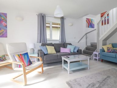 A living room with a gray sofa blue armchair wooden chairs white coffee table and staircase at Rainbow Lodge in St Tudy