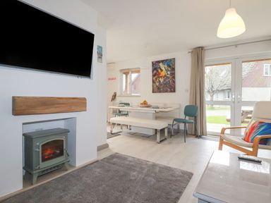 A living room with a wall mounted TV above a fireplace and a dining table with chairs and a bench at Rainbow Lodge in St Tudy