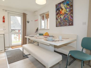 A dining area with a white table set with dishes and a fruit bowl with bananas a white bench and blue chairs a window and door with beige curtains and an abstract painting on the wall at Rainbow Lodge in St Tudy