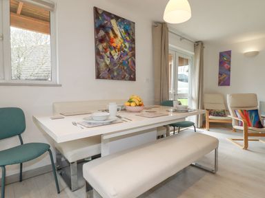 A dining area with a white table set with bowls plates cups and cutlery surrounded by chairs and benches with a fruit bowl on the table at Rainbow Lodge in St Tudy