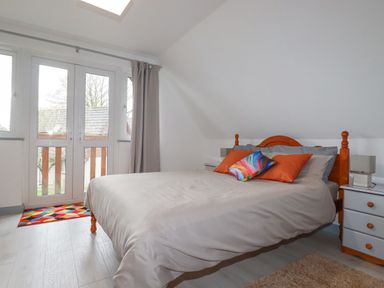 A bedroom with a wooden bed frame a bed with pillows a bedside table with lamps and glass doors with curtains at Rainbow Lodge in St Tudy