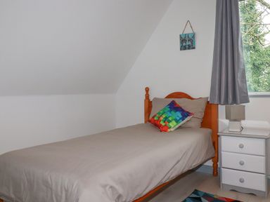 A bedroom with a single wooden bed with gray bedding and a colorful cushion next to a window with gray curtains and a white bedside table with a lamp at Rainbow Lodge in St Tudy