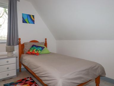 A bedroom with a single wooden bed a bedside table with a lamp a window with gray curtains and a colorful rug at Rainbow Lodge in St Tudy
