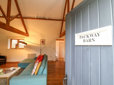 An open door with a sign reading Packway Barn overlooking a living room with a blue sofa and wooden beams at Packway Barn in Halesworth