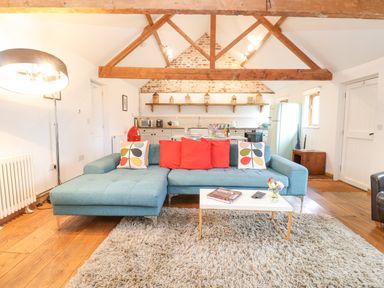 An open living area with a blue sectional sofa red and patterned pillows a white coffee table rug wooden floor and kitchen in the background at Packway Barn in Halesworth