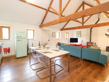 A combined kitchen and living area with a dining table, sofa, wood beams, and a stove at Packway Barn in Halesworth