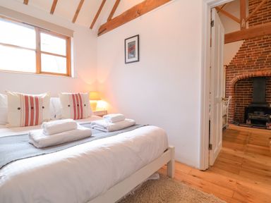 A bedroom with a bed white bedding and pillows a window wooden beams and a door opening to a room with a brick fireplace at Packway Barn in Halesworth