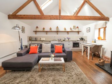 A living room with a gray sectional sofa orange pillows a coffee table and a kitchen with white cabinets and wooden beams at The Nook in Chediston near Halesworth