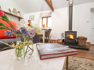 A glass vase with flowers and a book on a table in a living room with a wood stove and baskets at The Nook in Chediston near Halesworth