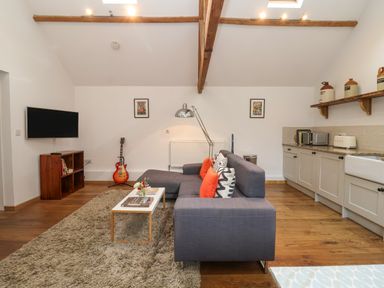 A living room with a gray sectional sofa and orange pillows a coffee table a guitar mounted tv and kitchen cabinets at The Nook in Chediston near Halesworth