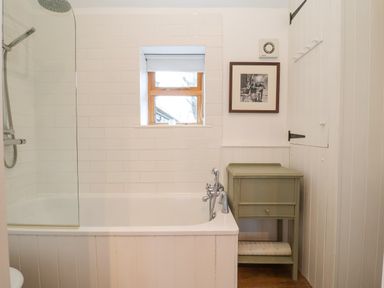 A bathroom with a bathtub shower combination a small window a framed picture and a green wooden side table at The Nook in Chediston near Halesworth