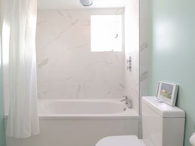 A bathroom with a bathtub a shower curtain a small window and a toilet with a framed picture on top at Twosome in Chediston near Halesworth