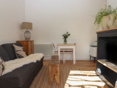 A living room with a dark sofa, wooden bench, small dining table with chairs, lamp on a cabinet and plants on a black shelf at The Apple Shed in Upexe near Silverton