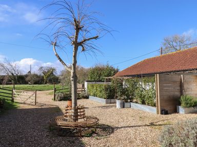 An outdoor area with a tree surrounded by a circular metal bench a gravel path hedges a wooden gate and a building with a red roof at The Apple Shed in Upexe near Silverton