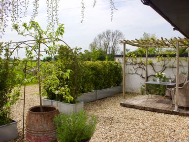 An outdoor garden area with gravel ground, potted plants, a wooden pergola, a table with two chairs and a wall with climbing plants at The Apple Shed in Upexe near Silverton