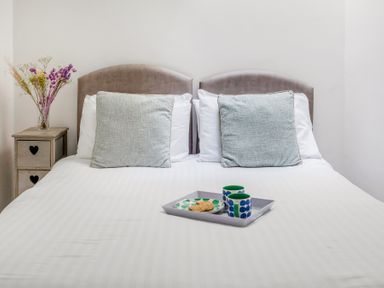 A bed with two pillows a tray with two mugs and a plate with cookies and a bedside table with flowers at The Cubby Hole in Sennen Cove