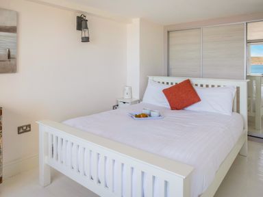 A bedroom with a white wooden bed with white bedding and an orange cushion and a tray with cups and food on the bed with sliding closet doors and a window overlooking water at The Cubby Hole in Sennen Cove