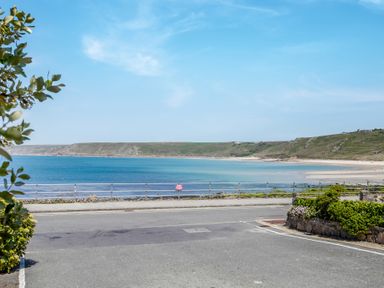 A coastal view with a road, stone wall, plants, and a beach with hills in the background at The Cubby Hole in Sennen Cove